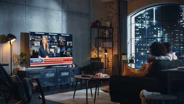 Black Couple Watches Evening News On TV While Sitting On A Couch At Home In The Night. Two Smart People Politely And In Civil Manner Discuss Important Political Issues Of The Day. Back View.