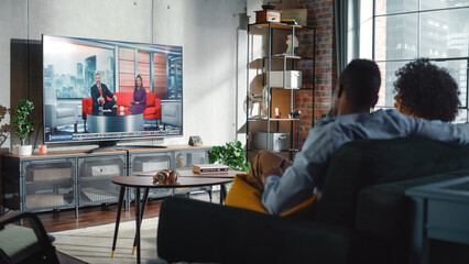 Boyfriend and Girlfriend Watching News Show on TV While Sitting on a Couch at Home on the Weekend. Two Hosts Talk and Joke on Televison. Cozy Living Room with Loft Interior Concept.