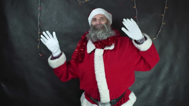 Santa claus is dancing on dark gray background. Strange middle-aged man with gray beard in Christmas costume is fooling around