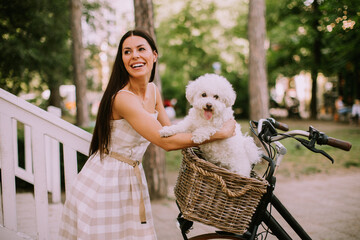 Young woman putting white bichon frise dog in the basket of electric bike - obrazy, fototapety, plakaty