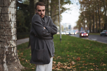 Depressive looking young man in dark coat standing on the street with lots of cars with headlights on going in background. Crisis and unemployment concept.