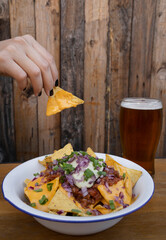 Eating nachos. Closeup view of a woman's hand holding a nacho dipped in cheddar cheese. A glass of beer besides the white bowl.