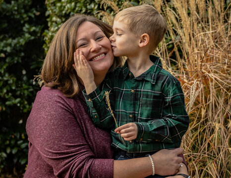 Mother and child posing and smiling while son kisses mom on the cheek softly with reeds and bushes in background sweet parent moment