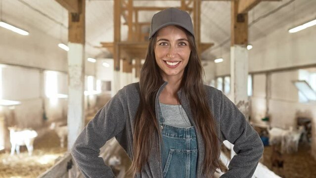 Close up of happy Caucasian beautiful female farmer in cap standing in animal farm in barn and smiling to camera. Joyful positive female shepherd indoors in livestock shed. Farming, goat farm concept