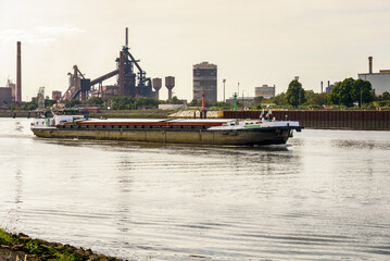 Cargo barge passing past a riverside steel plant while sailing up a river at sunset in summer