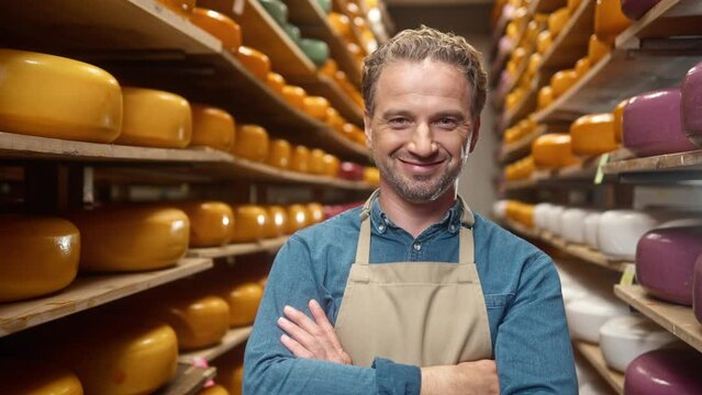 Close up of happy middle-aged Caucasian man in apron standing at storage house with cheese heads smiling to camera in own dairy factory. Wheels of cheese on wooden shelves. Craft production business