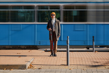 Blind man with a cane stopped on the sidewalk in front of a passing tram