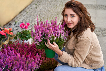 Young beautiful woman holding a pot with blooming violet heather plant in a street flower shop.