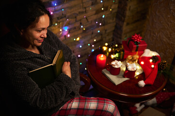 Delightful woman reads a book by candlelight, sitting at a table with Christmas gifts and garlands