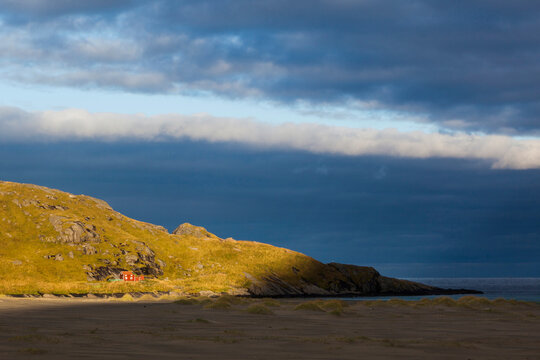 A shaft of morning light illuminates a lone cabin above Bunes Beach, Moskenesoya, Lofoten Islands, Norway.