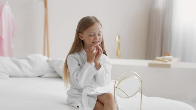 Adorable little girl wearing bathrobe touching her face, doing self massage, looking at mirror at bathroom, free space