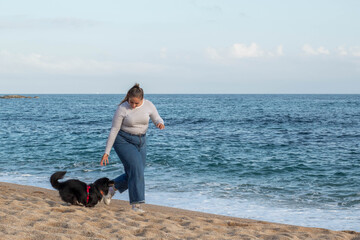 Mujer joven jugando en la playa con su bonito perro negro llamado Loki. Corriendo, sonriendo, queriéndose y abrazándose en actitud feliz y disfrutando de un agradable día - obrazy, fototapety, plakaty