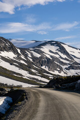 gray dirt road in the mountains in the summer when there is a lot of snow in the mountains on the gray stone rocks above which there are many blue skies with some thin fluffy clouds