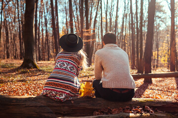 Back view of couple relaxing in autumn forest. Man and woman sitting on trunk enjoying fall landscape after walk