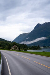 Naklejka premium a large gray asphalt road with a white line and a yellow solid line with a house with a fjord in the background and a mountain with a forest with white fog above it with clouds
