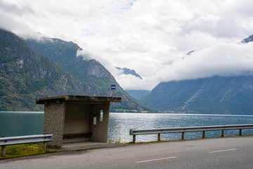 gray concrete bus stop on the side of the road behind which is a fjord with blue water and behind the fjord a beautiful view of big mountains covered with white fluffy clouds - Powered by Adobe
