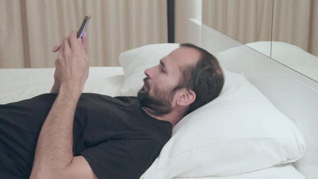 Man falls back on pillow, takes out his smartphone and begins to study news sites before going to bed. Smartphone mania, nomophobia. Mobile phone in bedroom.