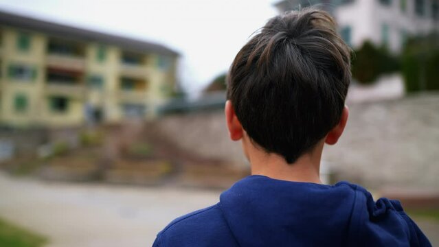 Tracking Shot Of Young Boy Standing Outside In City Park Looking At Camera With Serious Expression. Stoic Male Kid Turning Head