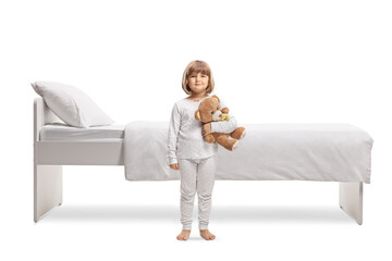 Full length portrait of a little girl in pajamas holding a teddy bear and standing in front of a bed - obrazy, fototapety, plakaty