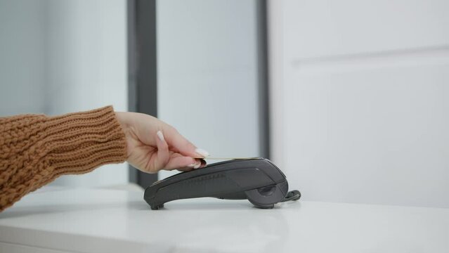 A female hand in a brown sweater applies a golden bank card to a terminal on a white neutral background. Wireless money transactions. Wireless payment.