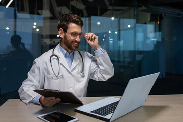 A young male doctor is talking to a patient using a video call from a laptop. He is sitting at a table in an office in a hospital. He holds a folder with documents, adjusts his glasses, talks, smiles.