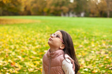 A little girl with long hair in glasses smiling looks up against the background of an autumn lawn