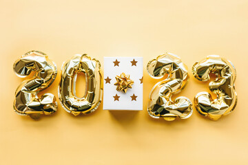 Balloons made of gold foil in the form of numbers 2023 and a gift box with a bow on a gold background. Celebrating Christmas, New Year and festive concept. Flat position, top view.