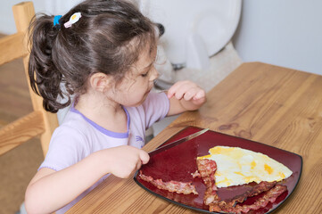 Young pretty girl eating eggs and bacon with knife and fork for the first time