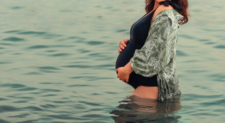 Motherhood and pregnancy. Young pregnant woman in swimsuit is standing in sea and holding her tummy. Copy space. Concept of water births
