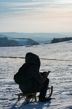 Rhoen Wasserkuppe in winter.  a man write sms messages on his phone sitting in the cold on top of a snowy mountain in winter on a children's sld wrapped in his winter warm jacket