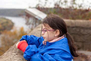 A beautiful little girl in glasses with a red bandage on her arm and in a raincoat looks into the distance in the autumn landscape