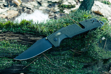 A sharp military knife with a black blade. Knife with a green handle on the moss in the forest.