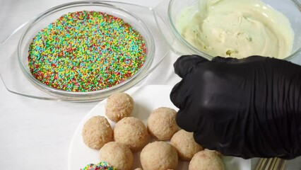 Preparation of desserts cake pops. Pastry chef at work. Proffessional baker in black gloves attending wooden stick to buiscuit ball in kitchen for swets. White chocolate and multicolored topping near 