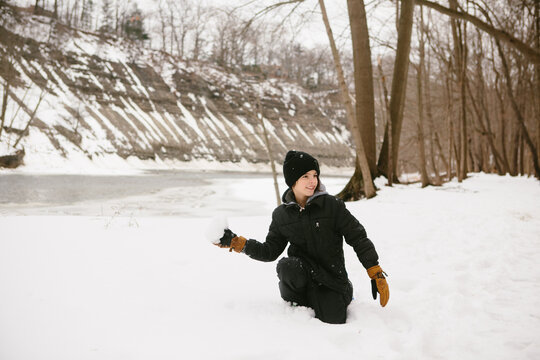 boy kneels in snowy wooded area and holds a snowball