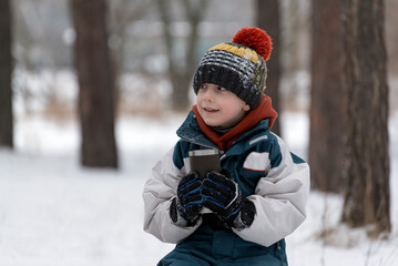 Fototapeta na wymiar Male child in knitted hat with hot beverage in thermos cup. Boy in snowy winter park.