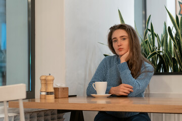 Cute young girl with brown hair in blue knitted sweater sits in cafe with cup of tea and looks thoughtfully into camera