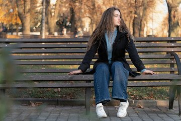 Teenage girl with long brown hair sits on bench in autumn park and waits for someone. Autumn time.