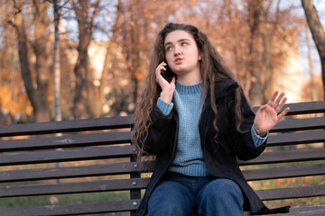 Young woman with long wavy brown hair sits on bench in park and indignaly talks on phone and gesticulating.