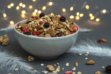 A bowl of muesli on a gray monochrome background, with white feathers and a side from a garland....