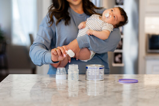 Mother Prepares Baby Formula In The Kitchen While Holding Infant