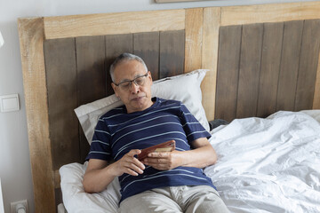 retired elderly man using smartphone at home, lying down on bed