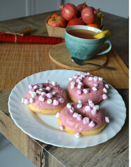 Cup of black tea with lemon and three donuts with pink icing on a plate on a wooden table. Sweet breakfast.