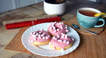 Cup of black tea with lemon and three donuts with pink icing on a plate on a wooden table. Sweet breakfast.