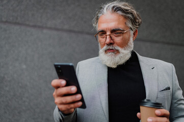 Senior business man standing outside while using smartphone and drinking coffee