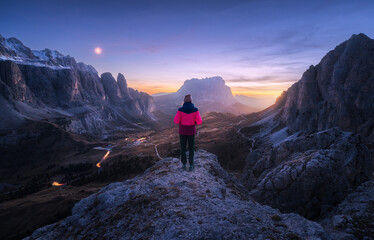 Woman on the rock and mountain peaks at night in autumn in Dolomites, Italy. Girl on the stone, high rocks, purple sky with moon, light trails on road at sunset in fall. Colorful landscape with cliffs