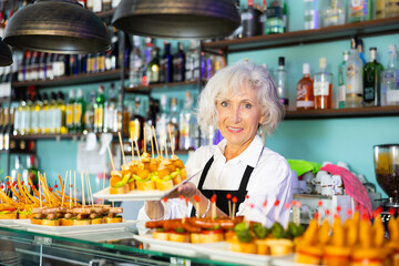 Positive mature female barman in uniform serving delicious pinchos at the counter of the bar. High quality photo