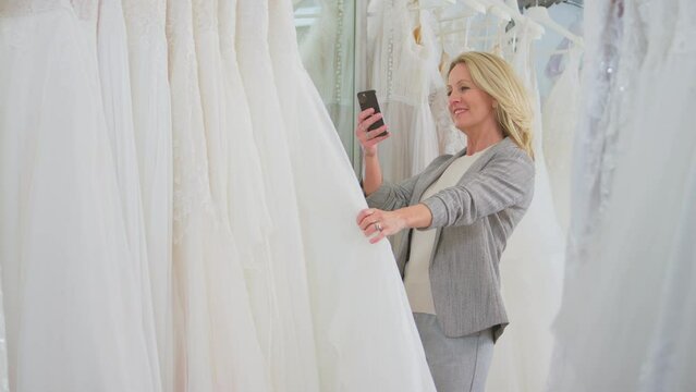 Mature woman in bridal store looking through wedding dresses taking picture on mobile phone - shot in slow motion