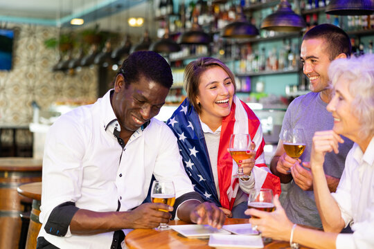 Young and senior football and soccer fans drinking beer at pub, cheering and celebrating scores. Girl with american flag on her shoulders - Powered by Adobe
