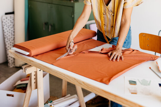 A standing unrecognizable woman cuts a piece of cloth on the table.
