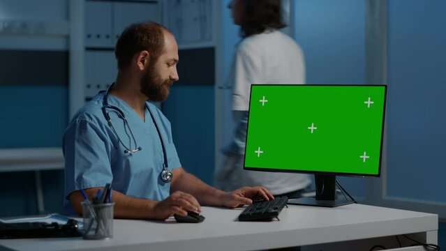 Practitioner man nurse sitting at desk typing patient illness report on computer with green screen chroma key mock up template. Physician assistant working over hours in hospital office.